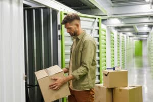 Side view at bearded adult man loading cardboard boxes into self storage unit.
