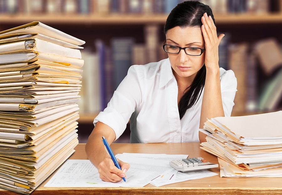 Stressed woman is working at a desk next to a tall stack of documents in file folders.