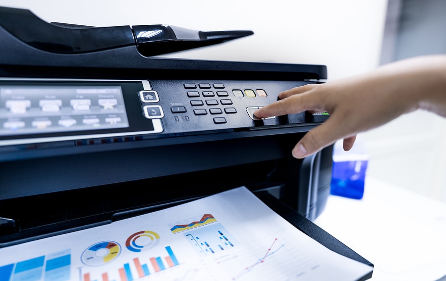 Hand pressing a button on an office printer as a page with colorful charts and graphs prints out.